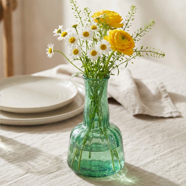 Strukturierte Glasvase in organischer Form mit Blumenstrauß aus Kamillen und Ranunkeln auf einem gedeckten Tisch.