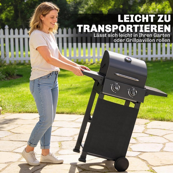 Eine Frau bewegt einen mobilen Gasgrill mit zwei Brennern auf Rollen über eine Steinterrasse in einem hellen Garten.