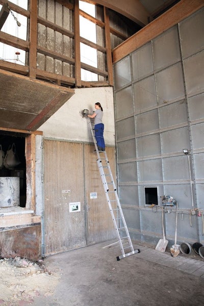 Eine Person steht auf einer hohen Aluminium Leiter in einer Scheune und montiert eine Außenleuchte an der Wand.