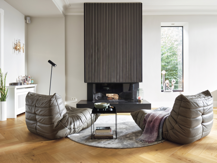 Modernes Wohnzimmer mit Kamin, dunkler Wandverkleidung aus Holzlamellen, zwei Leder-Sesseln und Parkettboden im Fischgrätmuster.