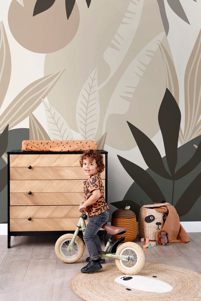 Kinderzimmer mit Kommode, Spielzeug und Kind auf einem Laufrad vor einer Fototapete mit Pflanzenmotiven.