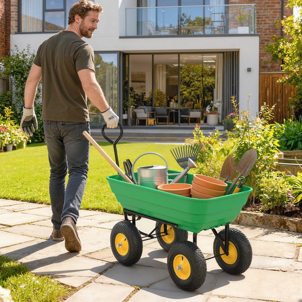 Mann zieht einen grünen Gartenwagen mit Gartenwerkzeugen, Gießkanne und Tontöpfen auf einem gepflasterten Weg in einem hellen, gepflegten Garten.
