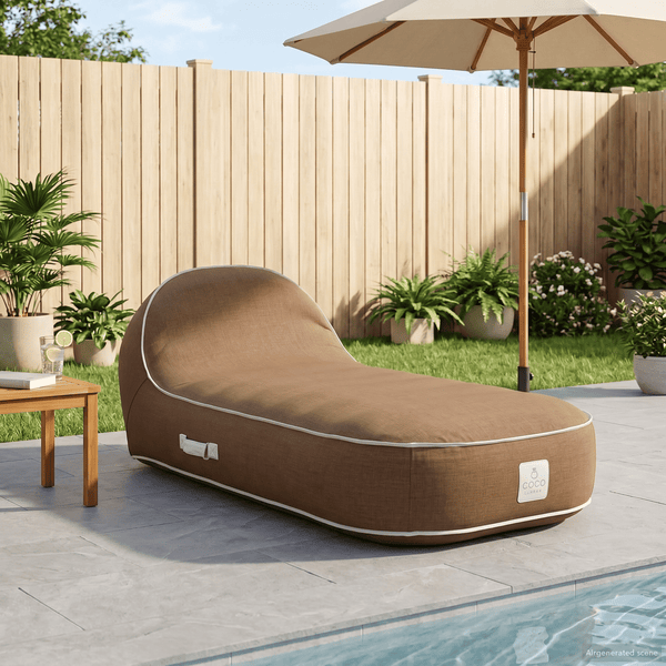Braune Gartenliege in Sitzsack-Form mit weißer Paspelierung auf einer Terrasse am Pool, unter einem Sonnenschirm mit Pflanzen im Hintergrund.