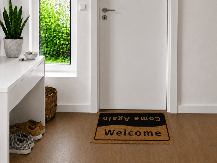 Rechteckige Fußmatte mit dem Schriftzug Welcome und Come Again vor einer weißen Tür in einem hellen Eingangsbereich mit Holzboden.