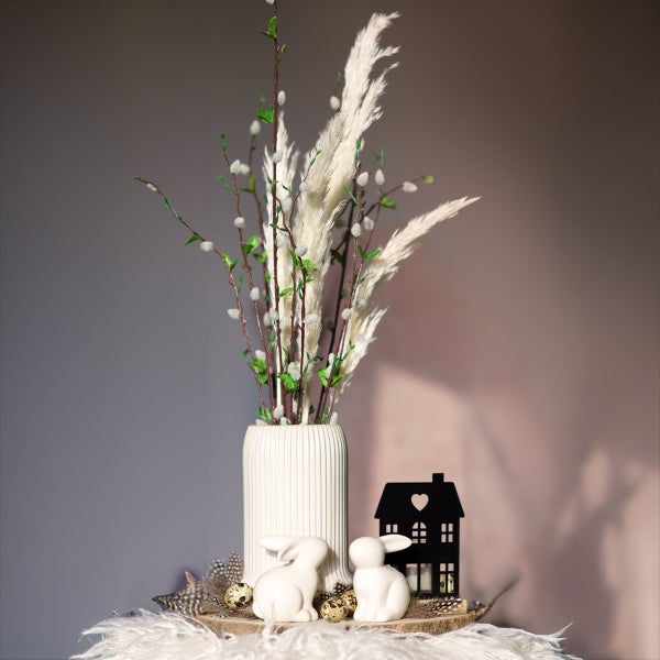 Osterdekoration auf Holzscheibe mit gerillter Vase, Pampasgras, Weidenkätzchen, weißen Keramikhasen und schwarzem Deko-Haus.