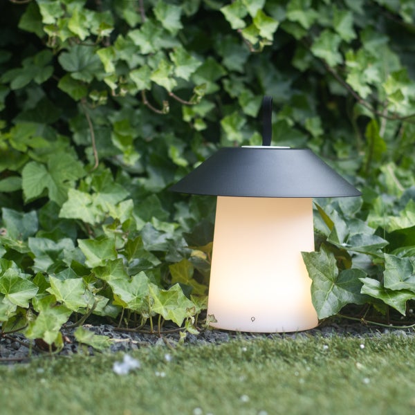 Tragbare Außenleuchte mit warmweißem Licht in einem Garten mit Efeu. Die Lampe hat einen dunklen Schirm und einen weißen, leuchtenden Korpus.