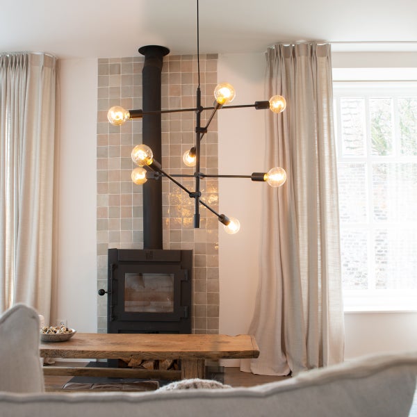 Wohnzimmer mit Kaminofen, Holztisch, Vorhängen und einer Hängelampe mit mehreren Glühbirnen.