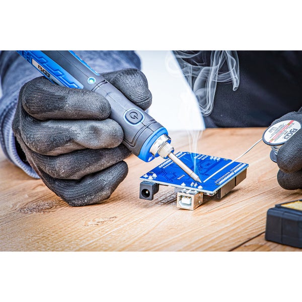 Person mit Schutzhandschuhen lötet mit einem Akku-Lötkolben Bauteile auf eine blaue Leiterplatte auf einem Holztisch.