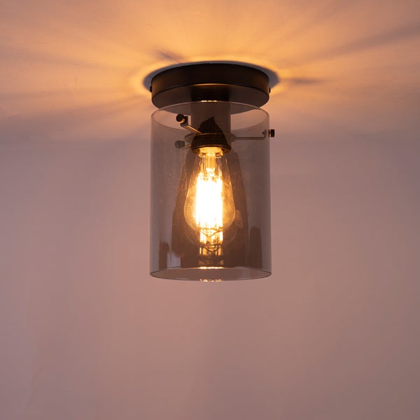 Zylindrische Deckenleuchte aus dunklem Glas und schwarzem Metall mit eingeschaltetem Filament-Leuchtmittel.