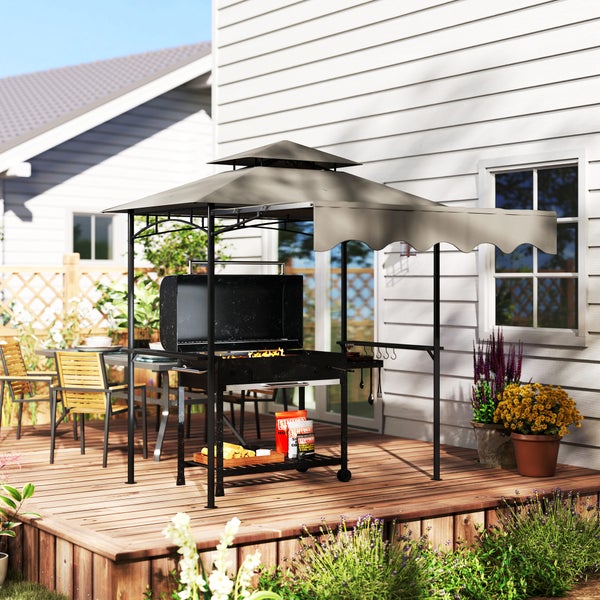 Grillpavillon mit Holzkohlegrill auf einer hellen Holzterrasse vor einem weißen Haus, umgeben von Gartenmöbeln und Blumen.