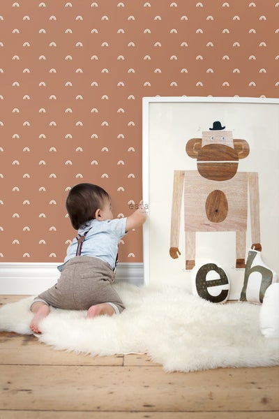 Ein Kind sitzt auf einem Schaffellteppich vor einer Wand mit Regenbogenmuster und einem Bild von einem Affen.