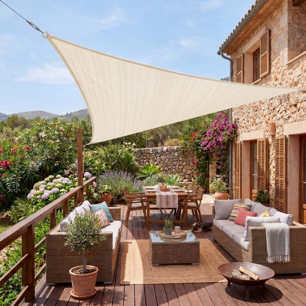 Dreieckiges helles Sonnensegel über einer Terrasse mit Gartenmöbeln, Esstisch und Pflanzen vor einem mediterranen Steinhaus.