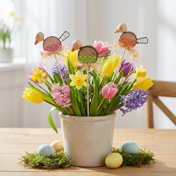Ostergesteck mit Frühlingsblumen, Holzsteckern in Vogelform und bemalten Eiern in einem Steinguttopf als Tischdekoration.