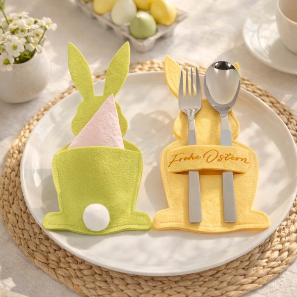 Helle Oster-Tischdekoration mit Besteckhaltern aus Filz in Hasenform auf einem Teller, Eiern und Blumen. Ein Halter trägt den Gruß Frohe Ostern.