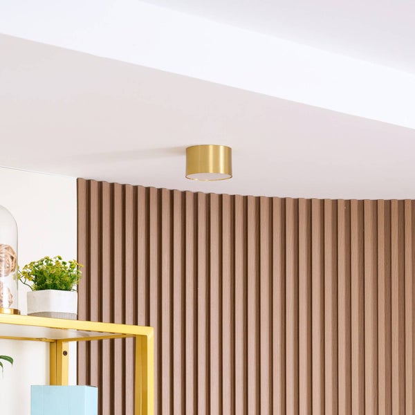 Zylindrische Deckenleuchte in Goldoptik an einer weißen Decke vor einer Wand mit vertikalen Holzlamellen.