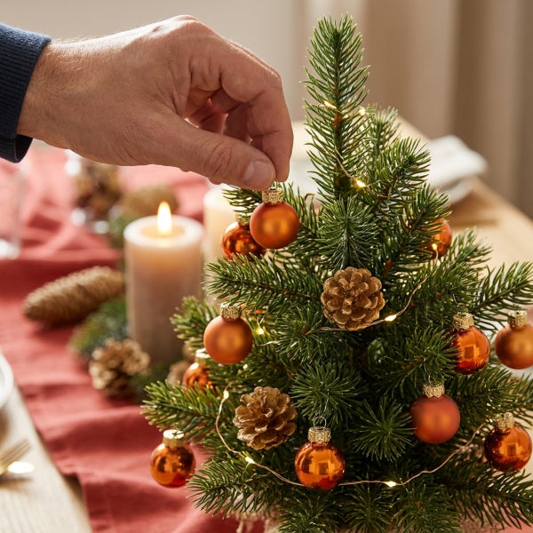 Hand dekoriert einen kleinen Weihnachtsbaum mit Kugeln und Zapfen, geschmückt mit einer Lichterkette auf einem festlich gedeckten Tisch.