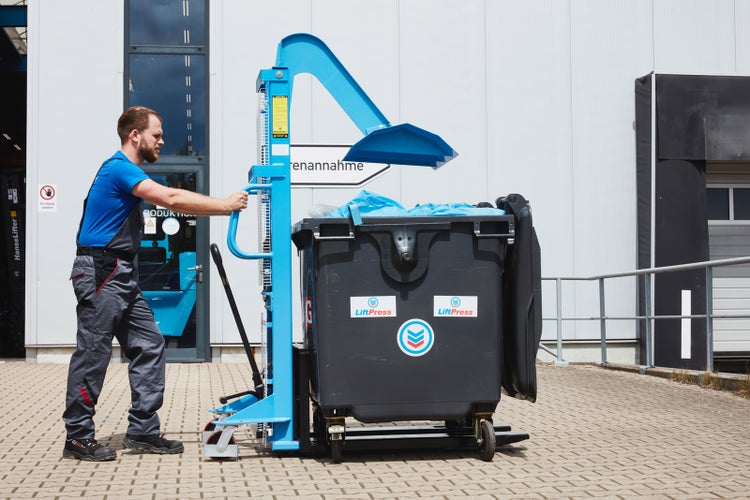 Ein Arbeiter bedient einen manuellen LiftPress Müllverdichter aus Metall an einem großen Abfallbehälter auf einem hellen, organisierten Industriehof.