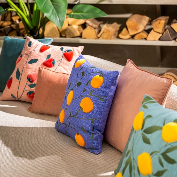 Dekorative Kissen mit Fruchtmotiven auf einem Sofa vor einem Regal mit Holzscheiten