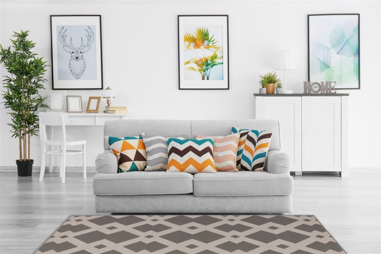 Modernes Wohnzimmer mit hellem Sofa, dekorativen Kissen mit geometrischen Mustern, Pflanzen, einem Teppich mit Rautenmuster und Wandbildern.