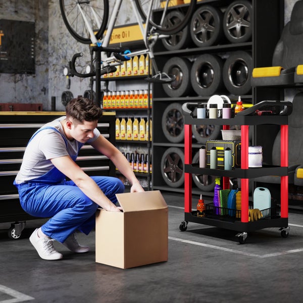 Handwerker in Arbeitskleidung öffnet einen Pappkarton in einer Werkstatt mit Werkstattwagen und Regalen mit Autozubehör.
