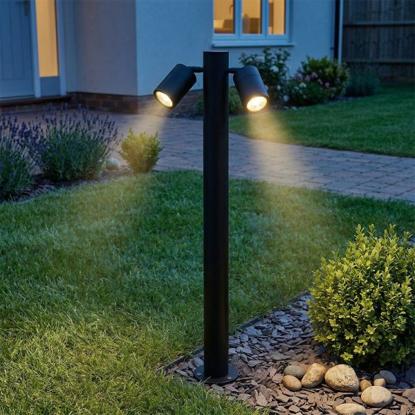 Schwarze Garten-Sockelleuchte mit zwei Strahlern beleuchtet einen Rasen und einen gepflasterten Weg in einem Garten bei Dämmerung.