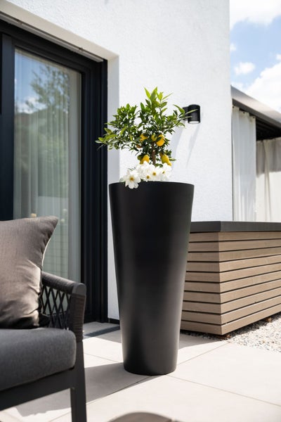 Ein hoher runder Pflanzkübel aus Kunststoff auf einer Terrasse neben einem Zitronenbaum und Sitzmöbeln vor einem modernen Wohnhaus.