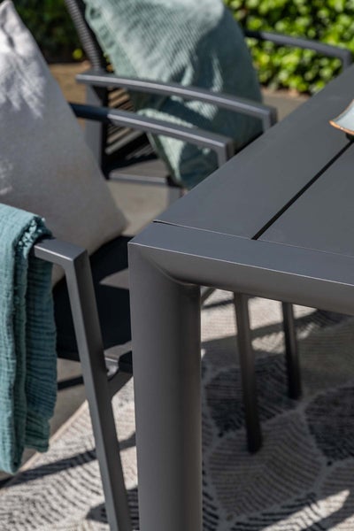 Detailansicht eines anthrazitfarbenen Gartentisches aus Metall mit Gartenstühlen, Kissen und einem Teppich im Außenbereich.