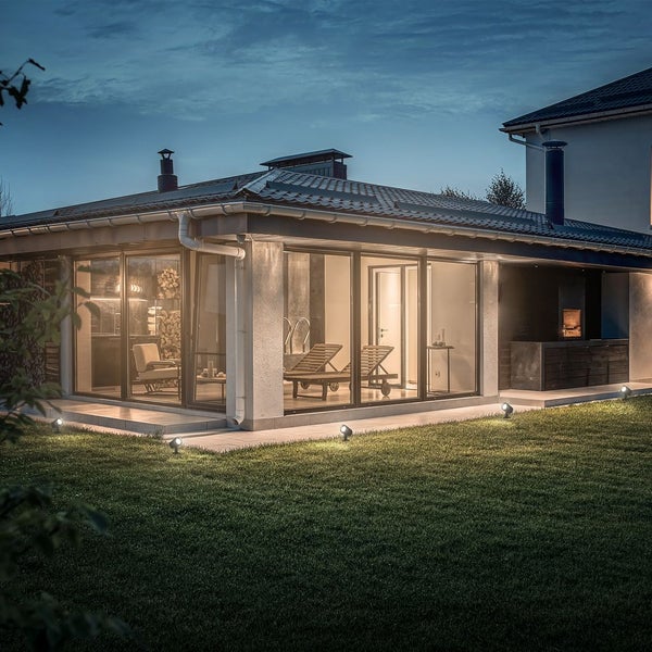 Modernes beleuchtetes Gartenhaus mit großen Glasfronten und Außenbeleuchtung durch Bodenstrahler auf einer Rasenfläche bei Nacht.