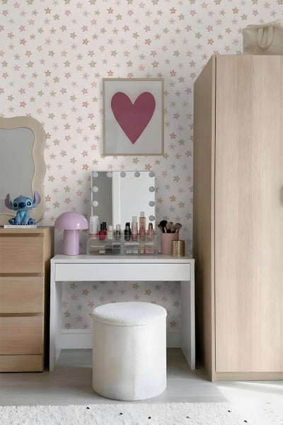 Ein Kinderzimmer mit Schminktisch, weißem Hocker und einer Tapete mit Blumenmuster vor einem hellen Holzschrank in einer organisierten Umgebung.