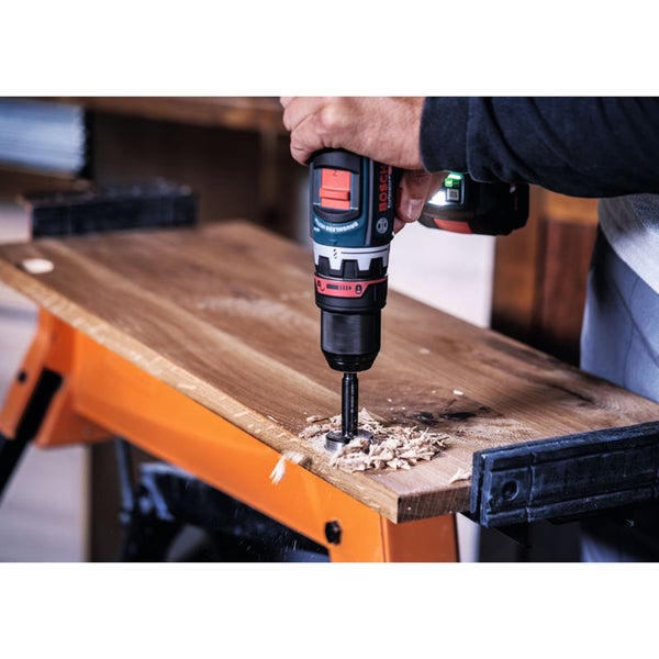 Eine Person bohrt mit einem Akku-Bohrschrauber und einem Forstnerbohrer ein Loch in eine hölzerne Waschtischplatte neben einem Waschbecken.