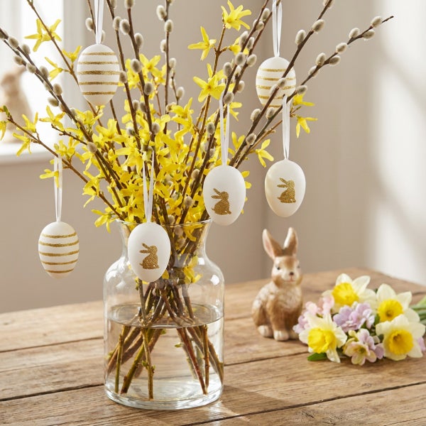 Osterstrauß aus Weidenkätzchen und Forsythien in einer Glasvase, dekoriert mit hängenden Ostereiern und einer Osterhasenfigur auf Holztisch.