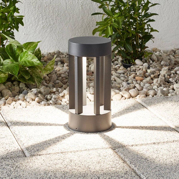 Moderne zylindrische LED-Sockelleuchte aus Metall in Anthrazit auf einer Terrasse mit Steinplatten, Kieselsteinen und Pflanzen.