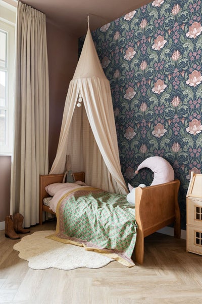 Kinderzimmer mit floral gemusterter Vliestapete, Holzbett mit textilem Betthimmel, langen Vorhängen und einem Puppenhaus aus Holz.