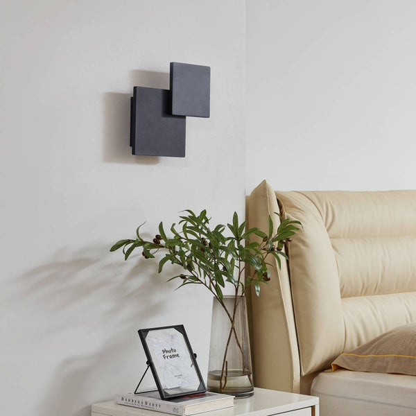 Moderne Wandleuchte aus zwei versetzten Quadraten über einem Nachttisch mit Vase und Bilderrahmen in einem hellen, organisierten Schlafzimmer.