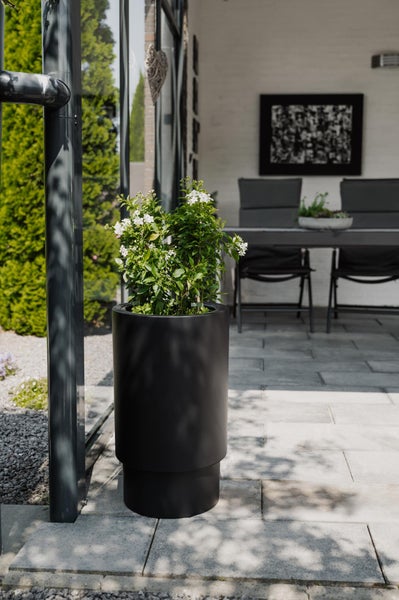 Hoher schwarzer Pflanzkübel mit grüner Pflanze und weißen Blüten auf einer gepflasterten Terrasse mit modernen Gartenmöbeln.