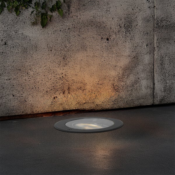 Runder Bodeneinbaustrahler im Außenbereich, bündig im Boden verbaut, beleuchtet eine Betonwand mit warmweißem Licht.