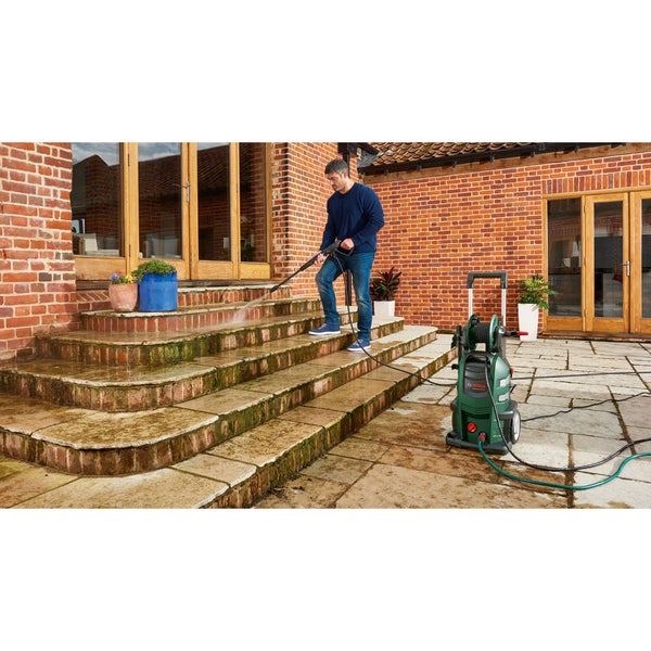Ein Mann reinigt mit einem Bosch Hochdruckreiniger bemooste Steinstufen im Außenbereich vor einem Backsteinhaus.