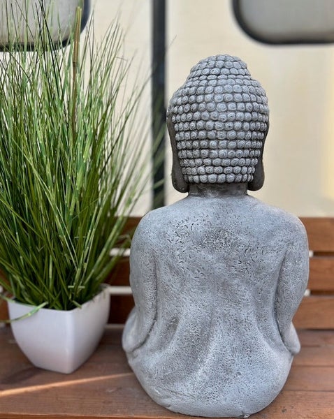 Sitzende Buddha-Figur in Steinoptik von hinten abgebildet auf einer Holzbank neben einer grünen Pflanze.