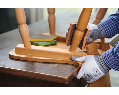 Eine Person schleift einen Stuhl mit einem Schleifschwamm. Auf dem Tisch liegen Schleifpads.