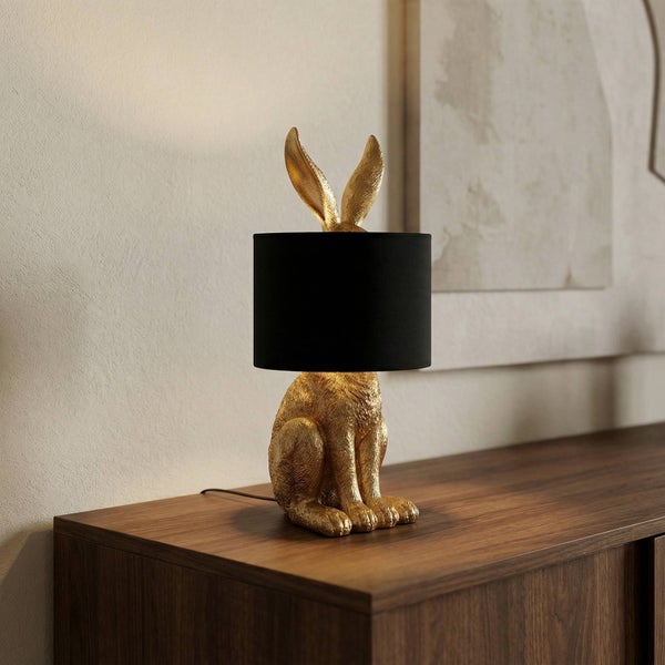 Tischleuchte in Form eines goldfarbenen Hasen mit schwarzem Stoffschirm auf einer Kommode aus Holz.