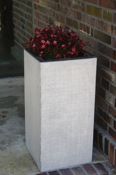 Hoher quadratischer Pflanzkübel in Betonoptik bepflanzt mit roten Blumen vor einer Ziegelwand.