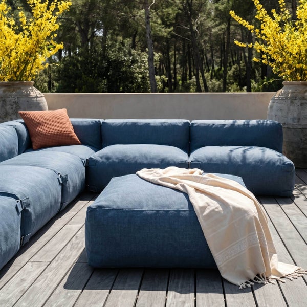 Modernes modulares Lounge-Sofa-Set mit Hocker auf einer Holzterrasse, blauer Stoffbezug, orangefarbenes Kissen und eine helle Decke.