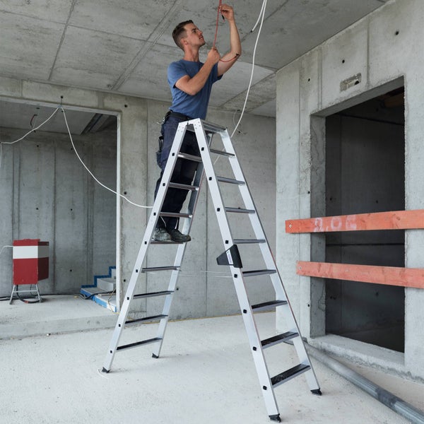 Ein Handwerker steht auf einer stabilen Aluminium Doppelstufenleiter bei Elektroinstallationsarbeiten auf einer Baustelle.