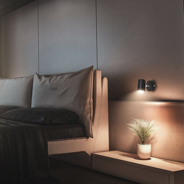 Schwarzer zylindrischer Wandstrahler in einer modernen Schlafzimmer-Szene. Das warme Licht beleuchtet eine kleine Pflanze und das Bettzeug.