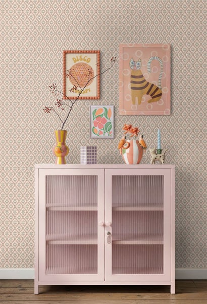 Wohnszene mit gemusterter Vliestapete und einem hellrosa Vitrinenschrank mit geriffelten Glastüren sowie Wandbildern.