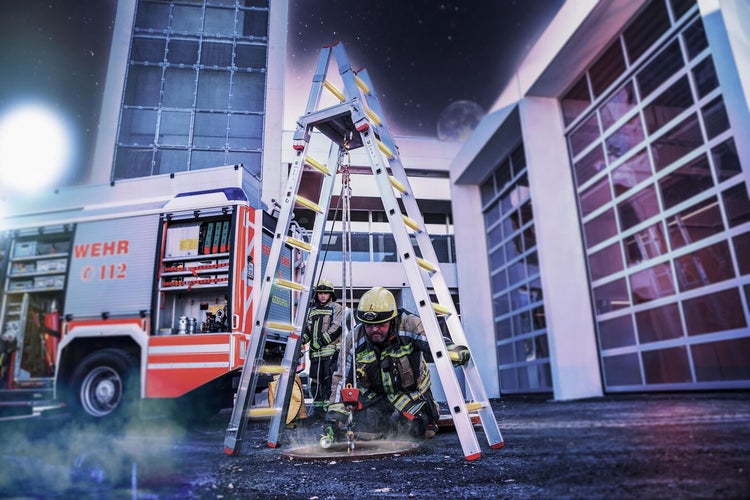 Feuerwehrleute führen eine Übung mit einer Aluminiumleiter und einem Feuerwehrfahrzeug im Hintergrund durch