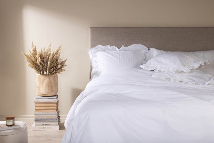 Hell gestaltetes Schlafzimmer mit weißer Bettwäsche, Kissen mit Rüschen, grauem Polsterkopfteil und Dekoration mit Trockenblumen.