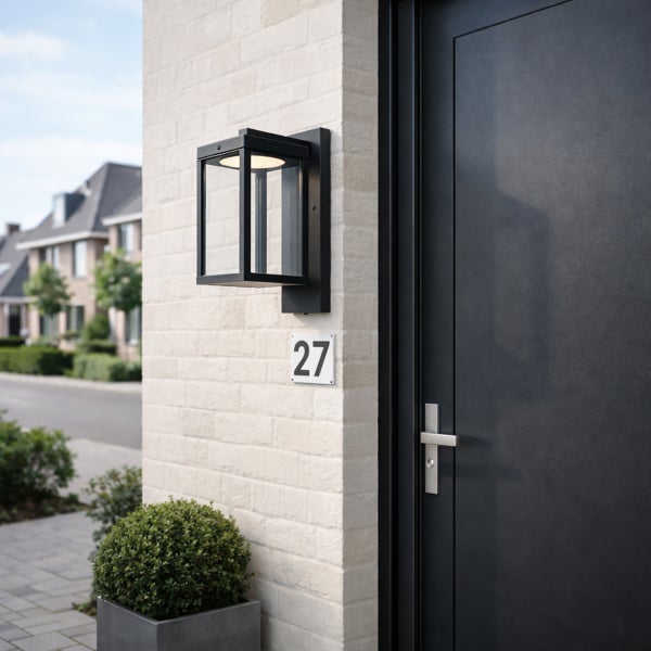 Moderne schwarze Außenwandleuchte mit Glaseinsätzen an einer hellen Klinkerwand neben einer dunklen Haustür.