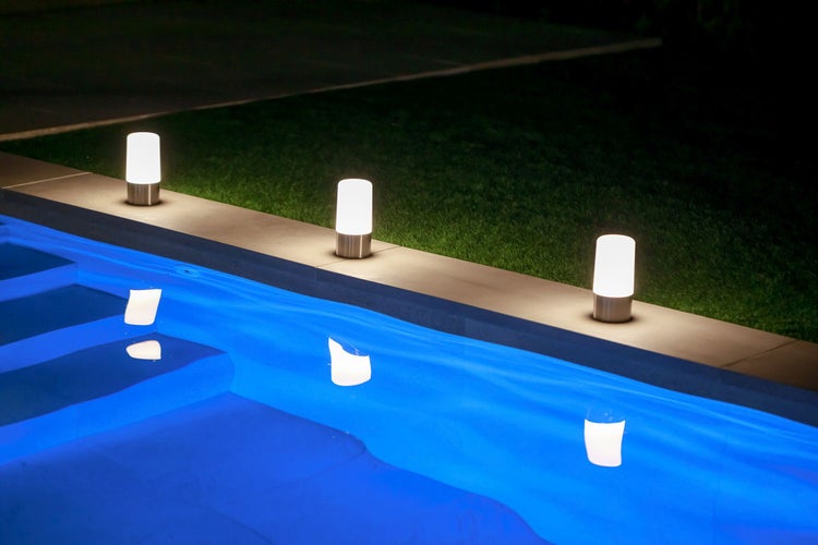 Drei moderne Außenleuchten in zylindrischer Form leuchten nachts den Rand eines Swimmingpools aus.
