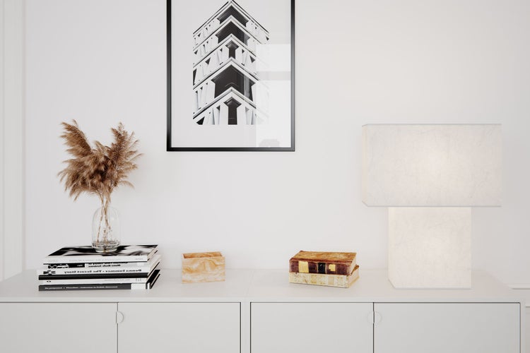 Dekoratives Arrangement auf einem weißen Sideboard mit einer Lampe, Büchern, einer Vase und einem gerahmten Bild.